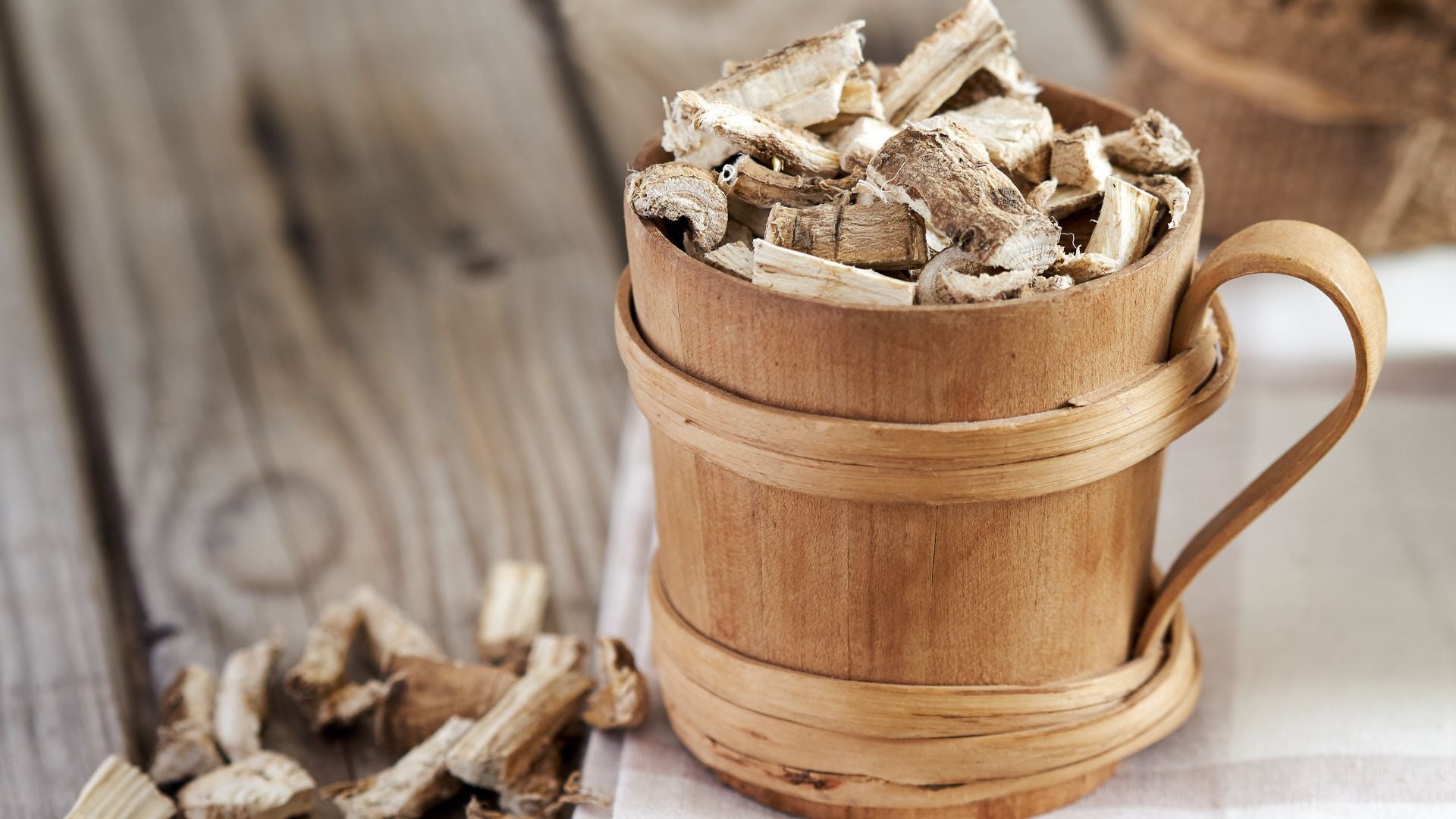 Une tasse en bois rustique débordant de racines séchées de Guimauve, posée sur un fond en bois avec des éclats de racines dispersés autour, représentant l'utilisation traditionnelle de la plante en phytothérapie.